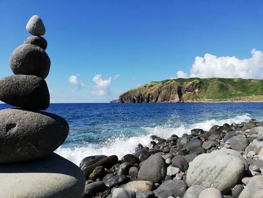 Stacked volcanic boulders on Valugan Boulder Beach with the Pacific coast and green cliffs beyond